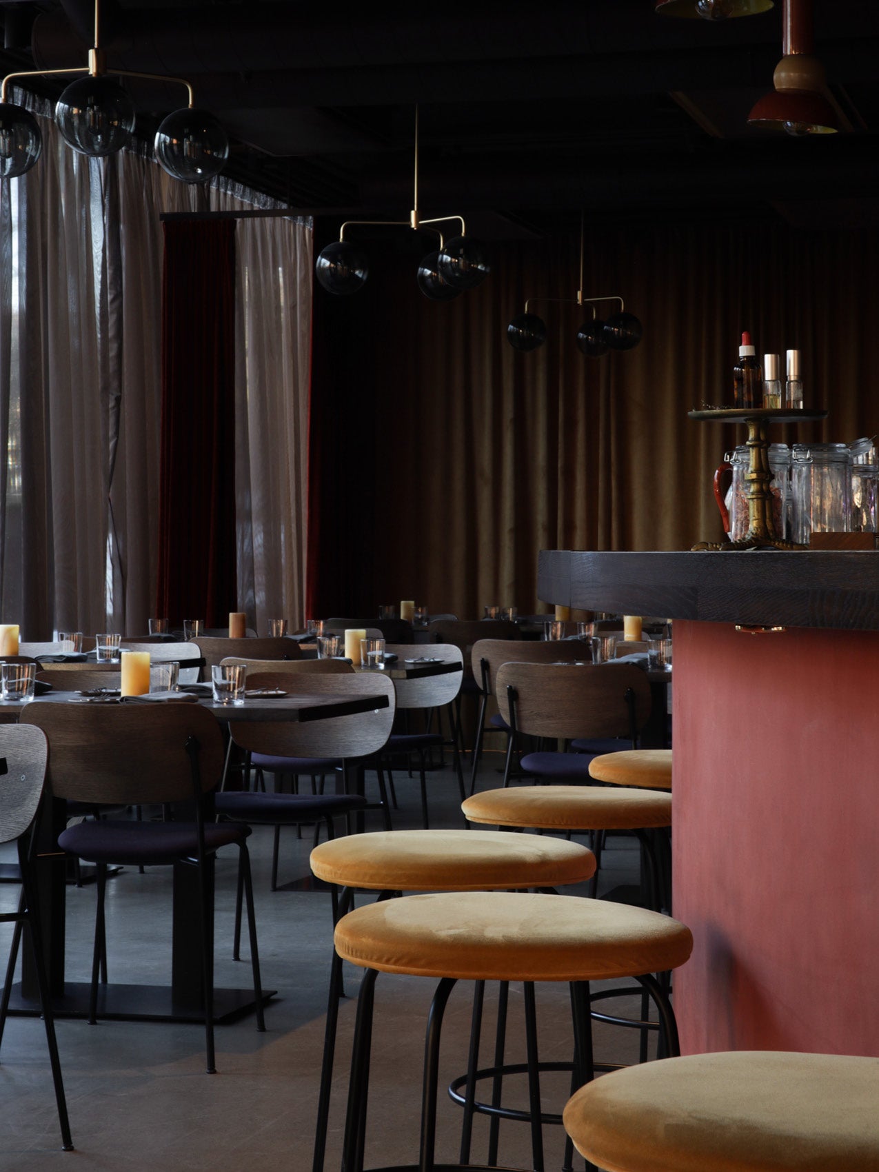 Restaurant interior with round mustard velvet bar stools, dark wood tables and chairs, smoked-glass pendant lights and lit