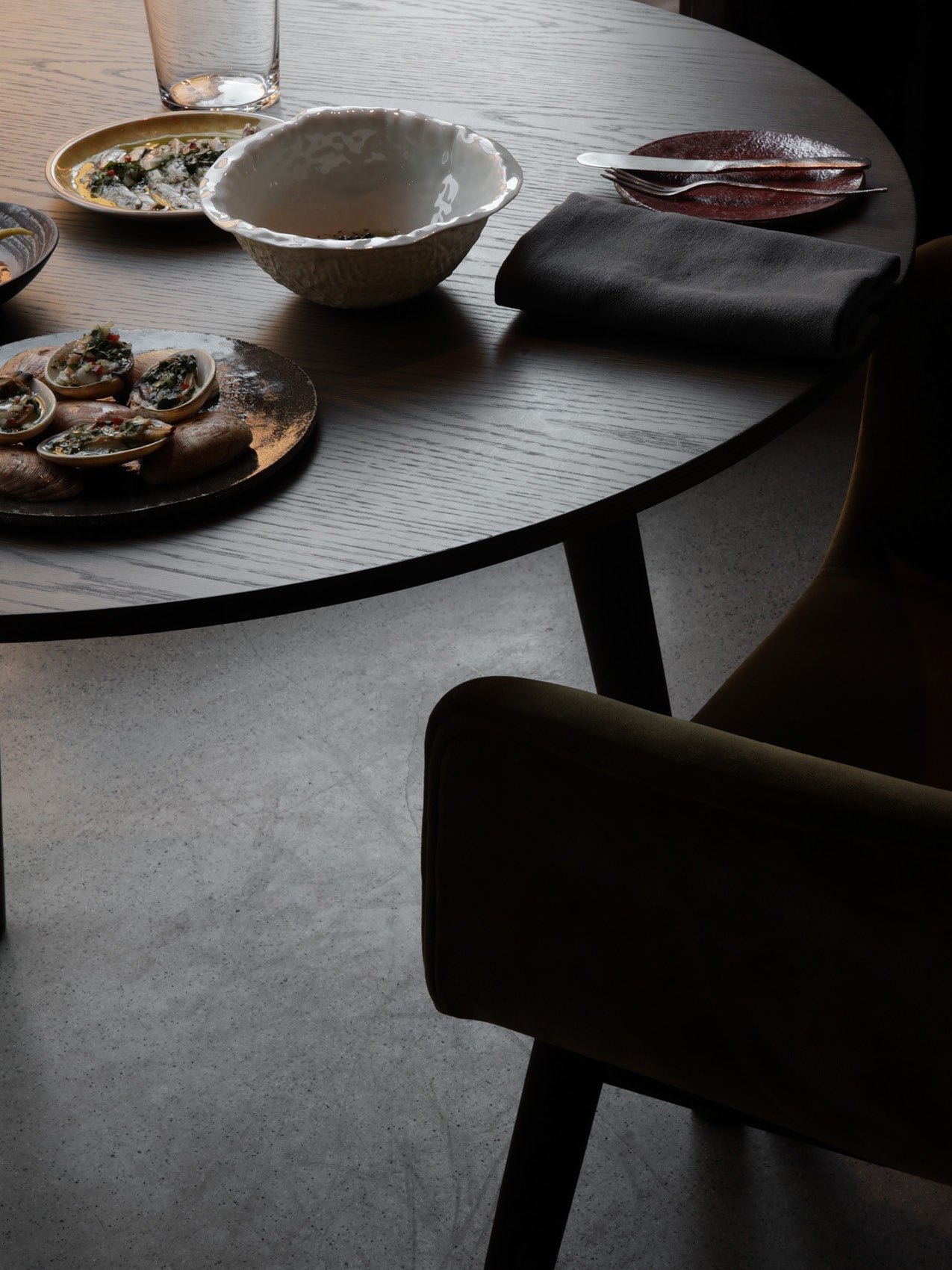 Dark wood round dining table with ceramic bowls, small plates of food, folded grey napkin and an upholstered chair.