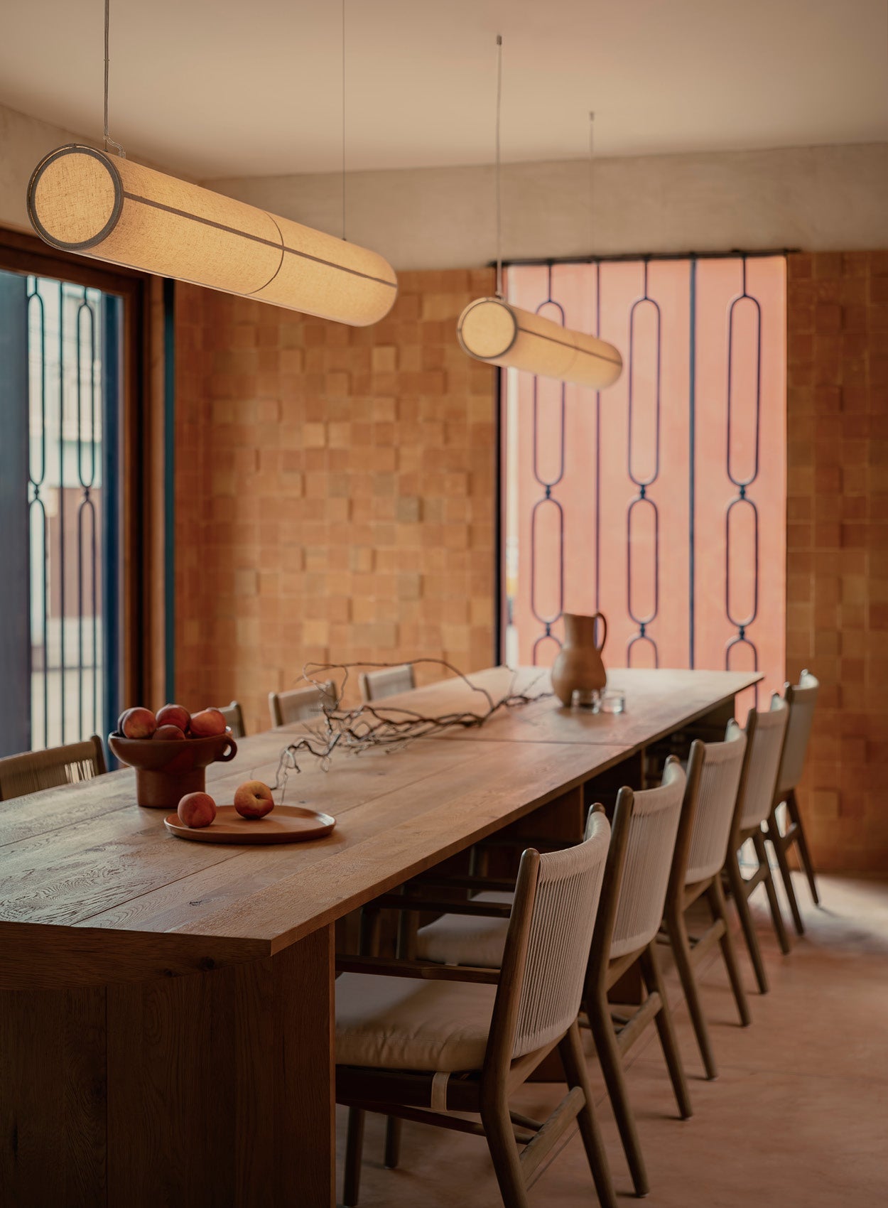 Long wooden dining table with woven-back chairs, two cylindrical beige fabric pendant lights and terracotta wall backdrop