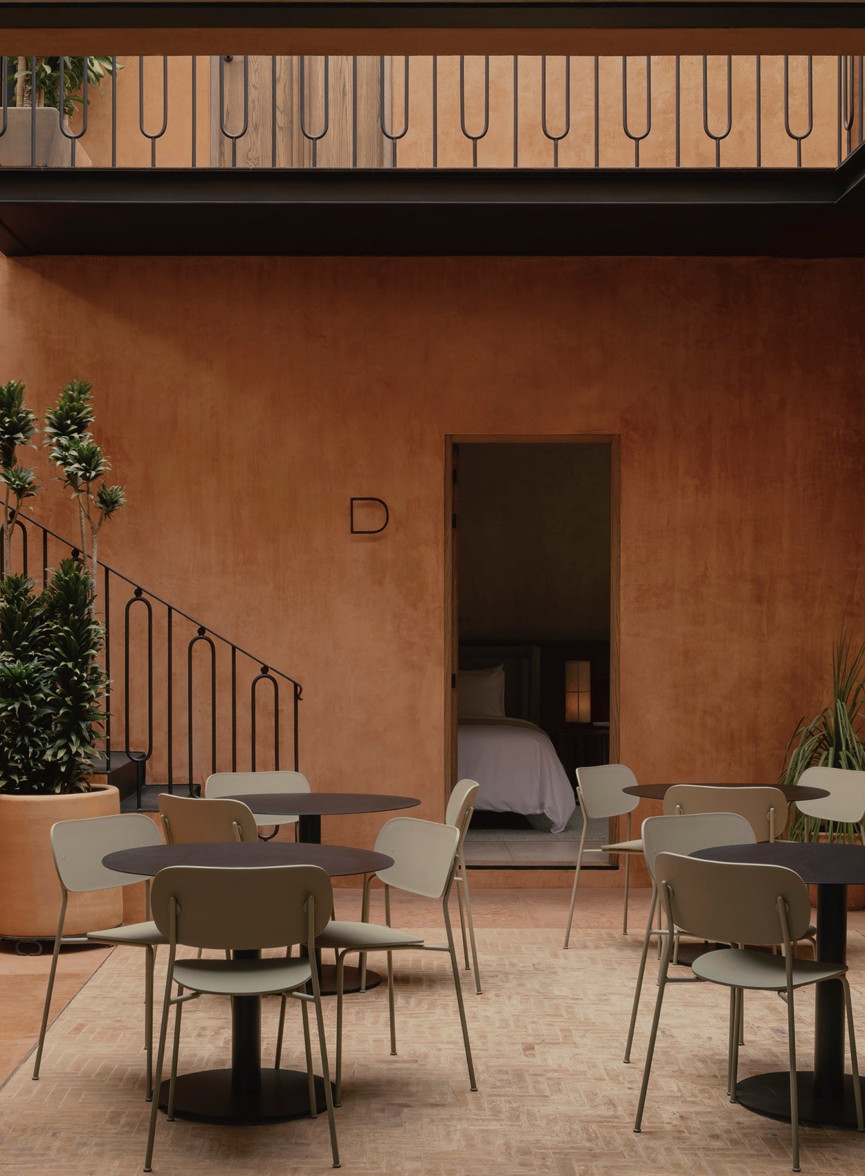 Courtyard dining area with olive-green metal chairs and round black tables against terracotta plaster walls.