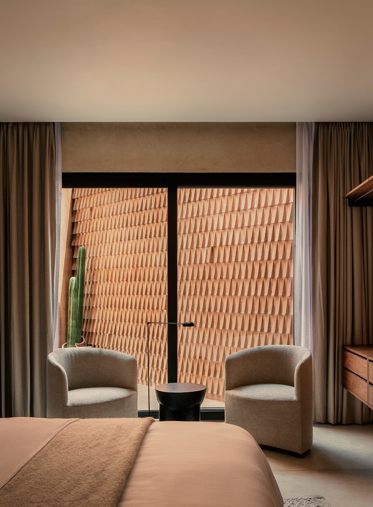 Minimal hotel seating area with two beige boucle armchairs, black round side table, tall cactus and textured terracotta wall