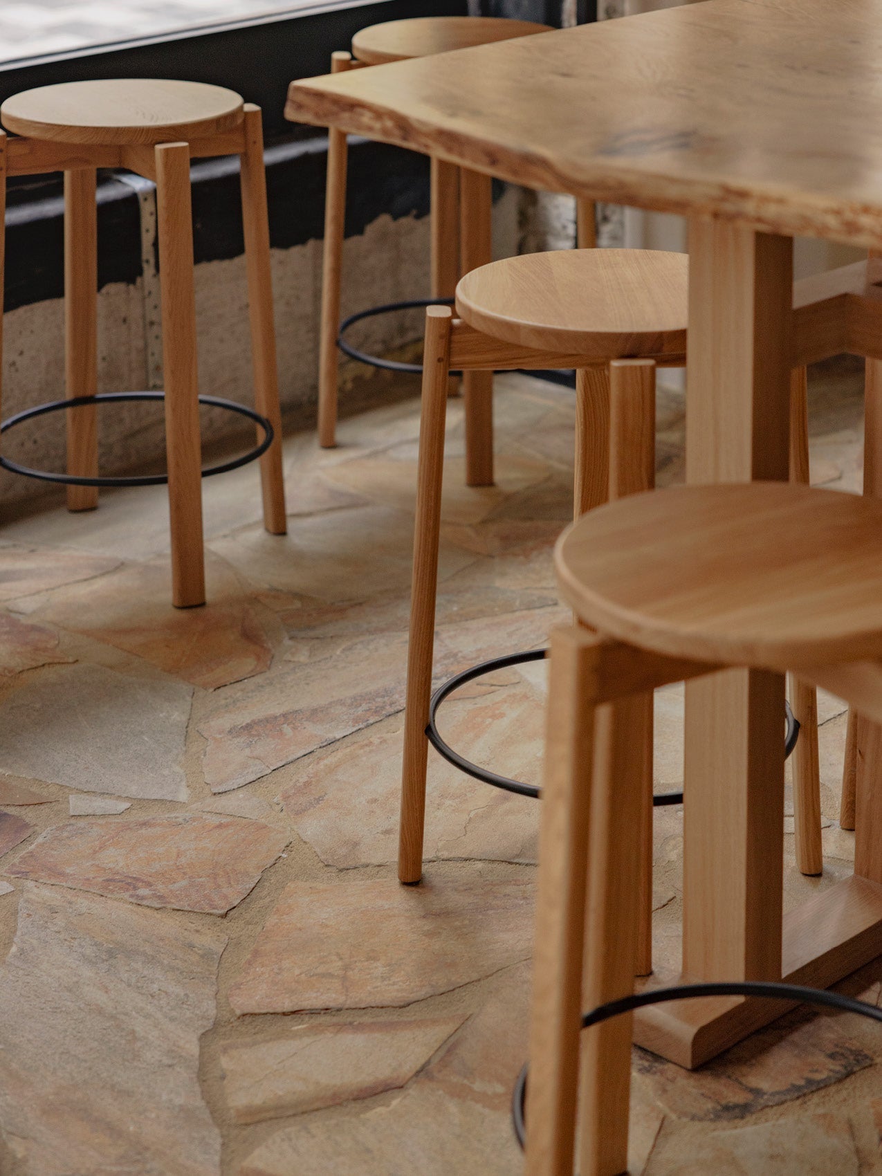 Natural oak round-seat bar stools with slim legs and black metal footrests beside a live-edge wooden table on stone floor