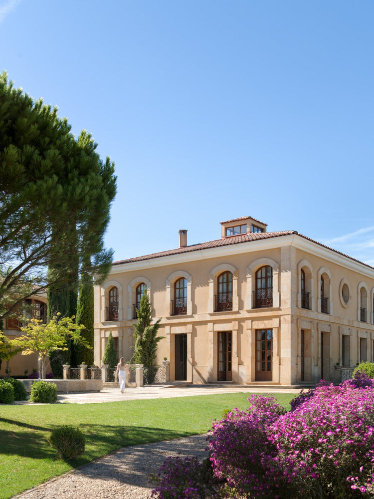 Mediterranean two-story beige stone villa with arched windows, terracotta roof, manicured lawn and purple flowering shrubs.