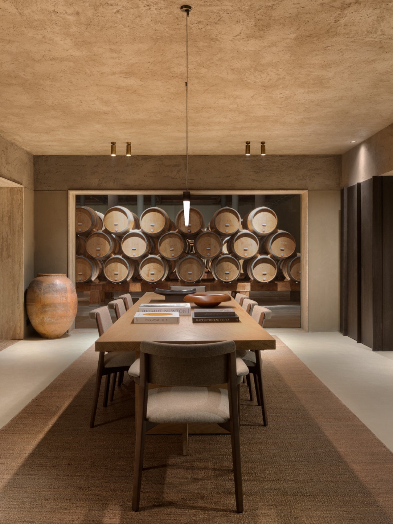 Minimal dining room with long oak table, upholstered chairs, pendant light and stacked wooden wine barrels behind glass.