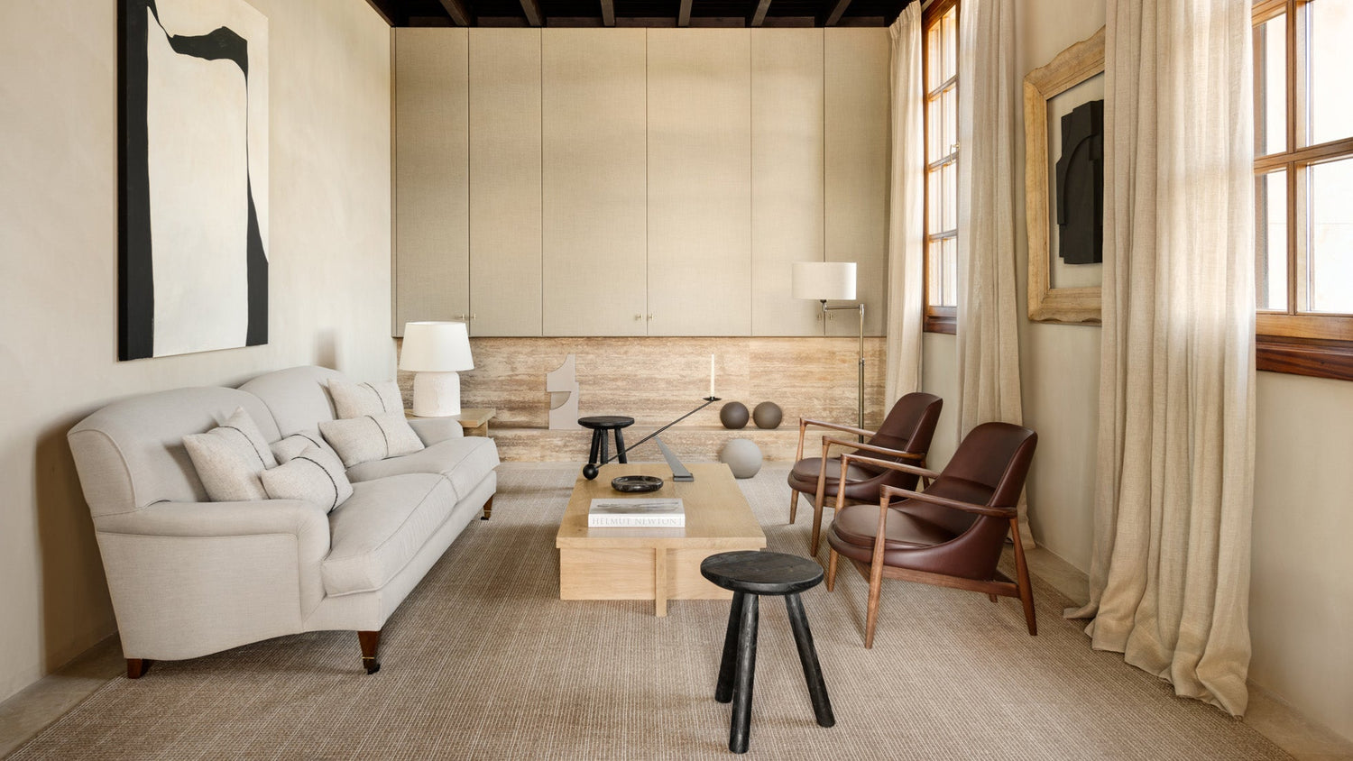 Neutral-toned living room with beige fabric sofa, wooden coffee table, black stools and two brown leather armchairs