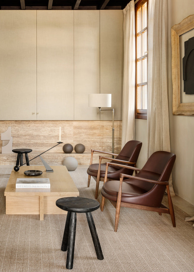 Minimal living room with two brown leather wooden-frame armchairs, light oak coffee table, black stools and floor lamp.