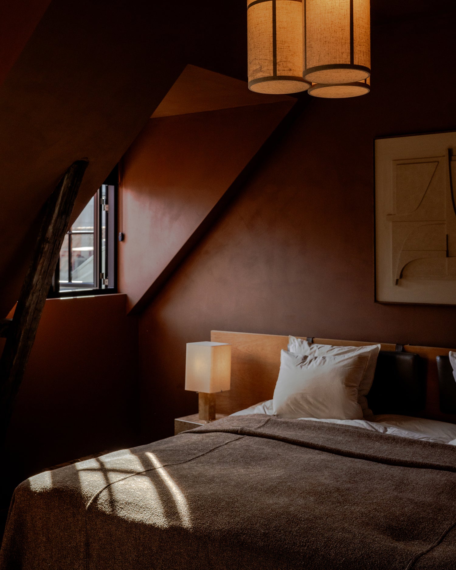 Cozy bedroom corner with brown textured bedcover, wooden headboard, cream pillows, cube bedside lamp and hanging fabric