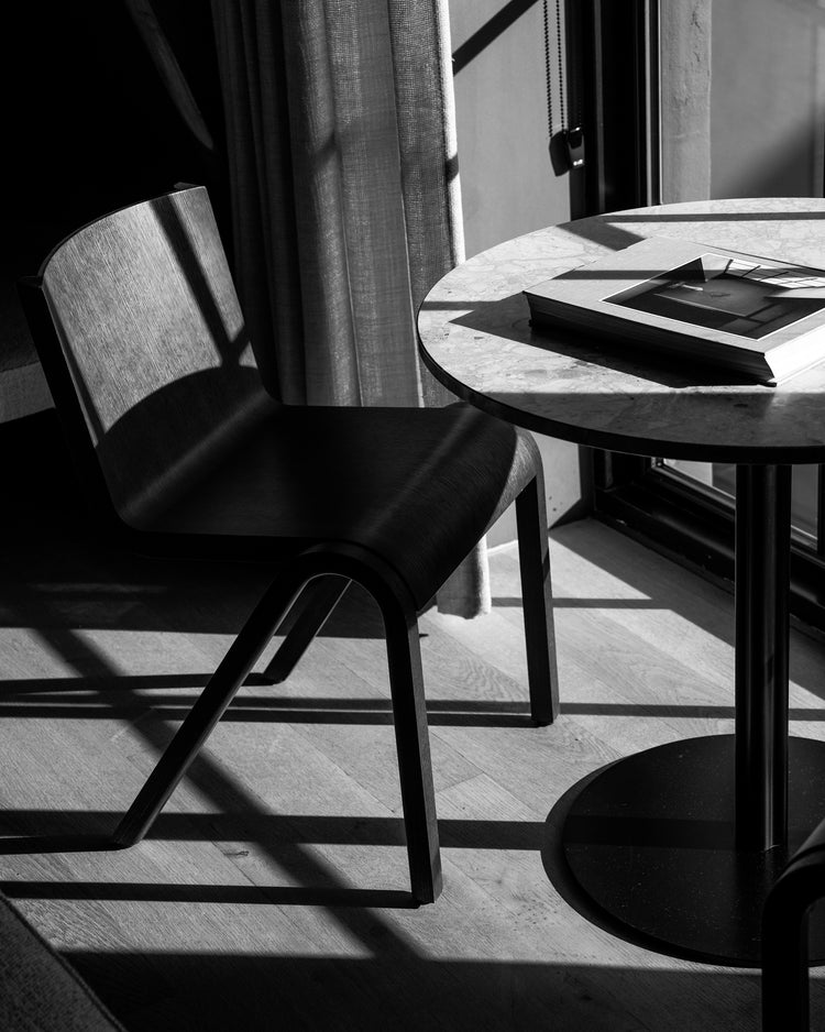 Black wooden chair beside round marble-topped table with a book, sunlight casting geometric shadows on wood floor