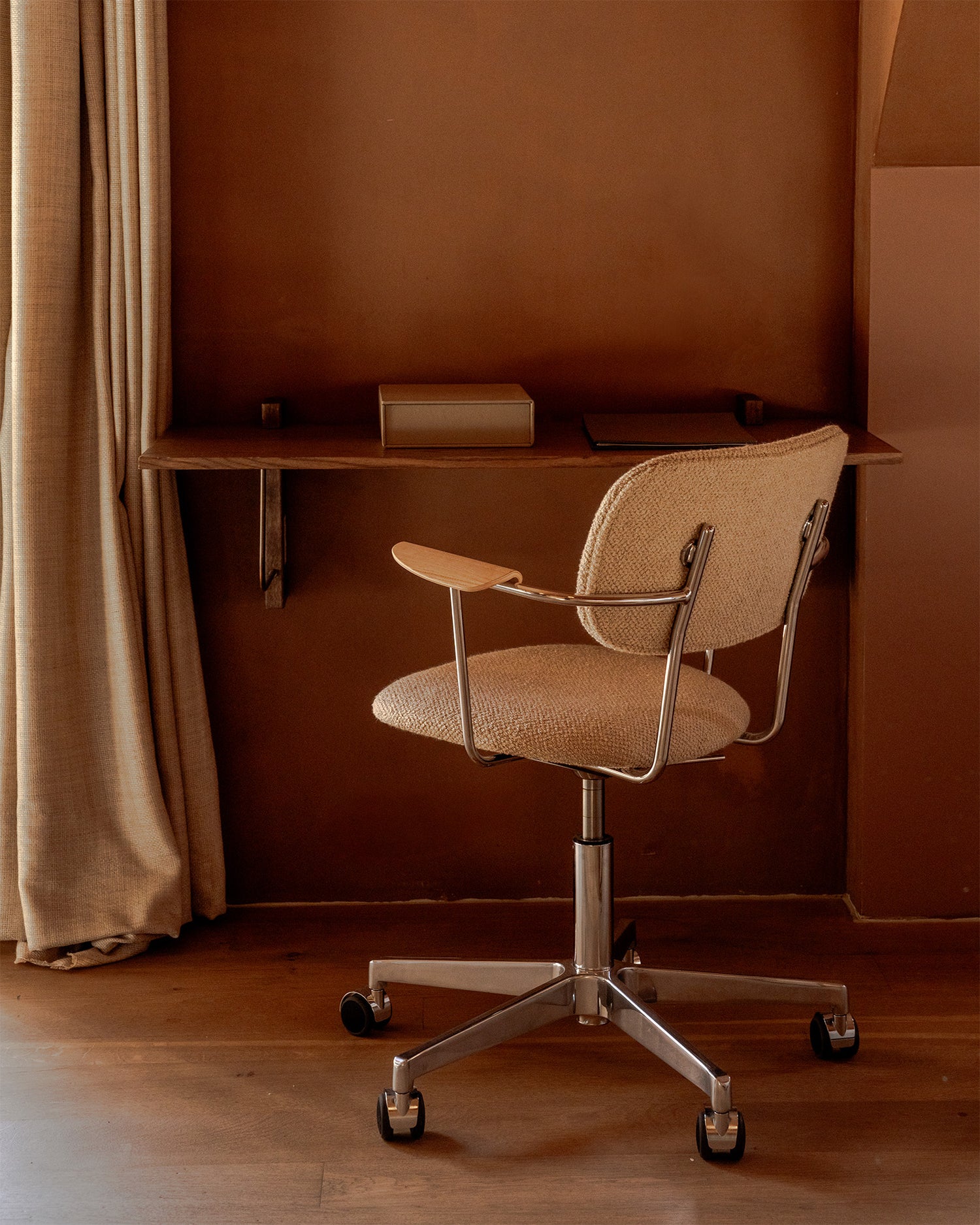 Beige boucle office chair with chrome swivel base and wooden armrest in front of a narrow wooden wall shelf and brown wall