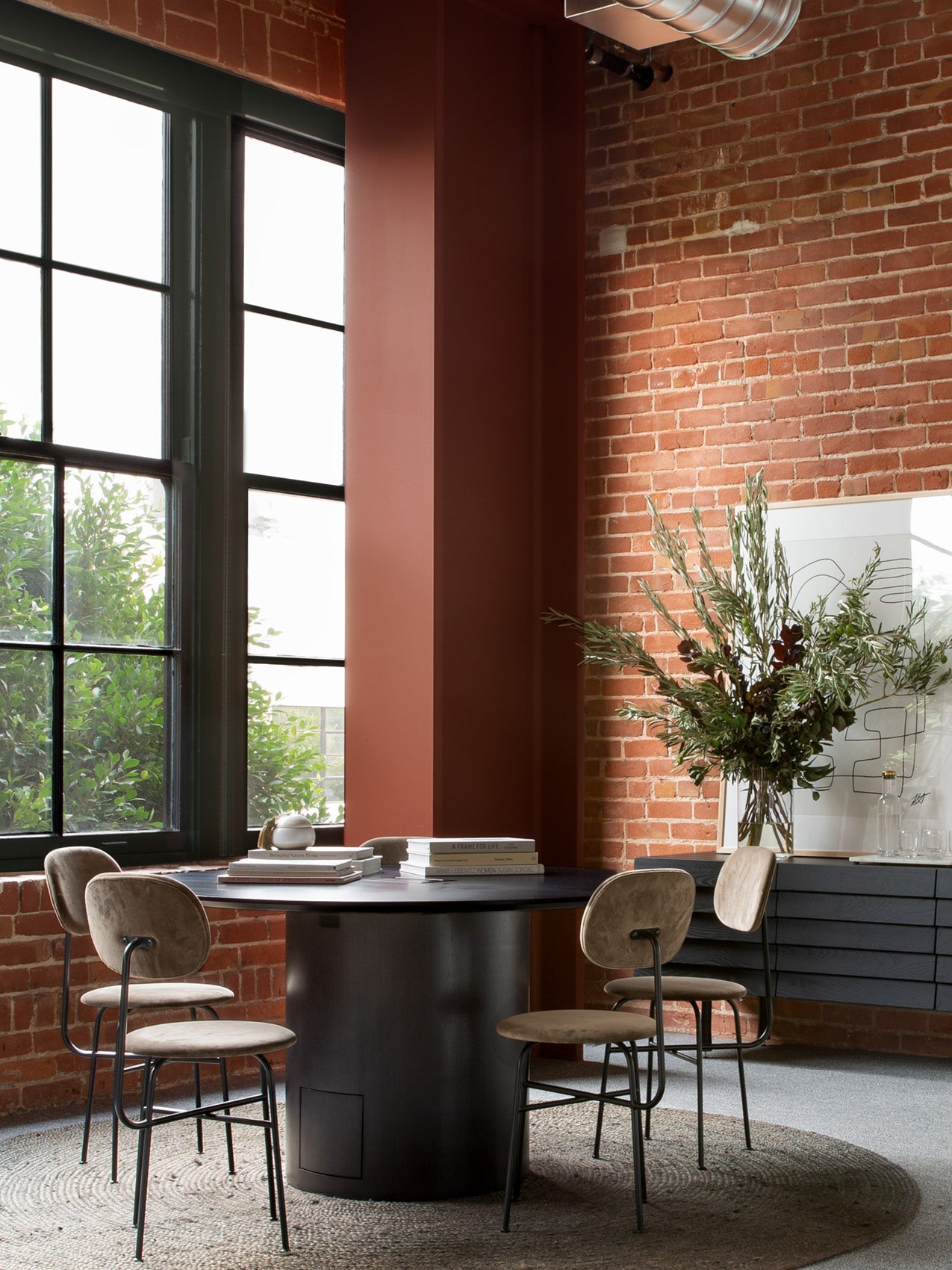 Round black dining table with suede beige bar stools, brick wall, large window and vase of greenery