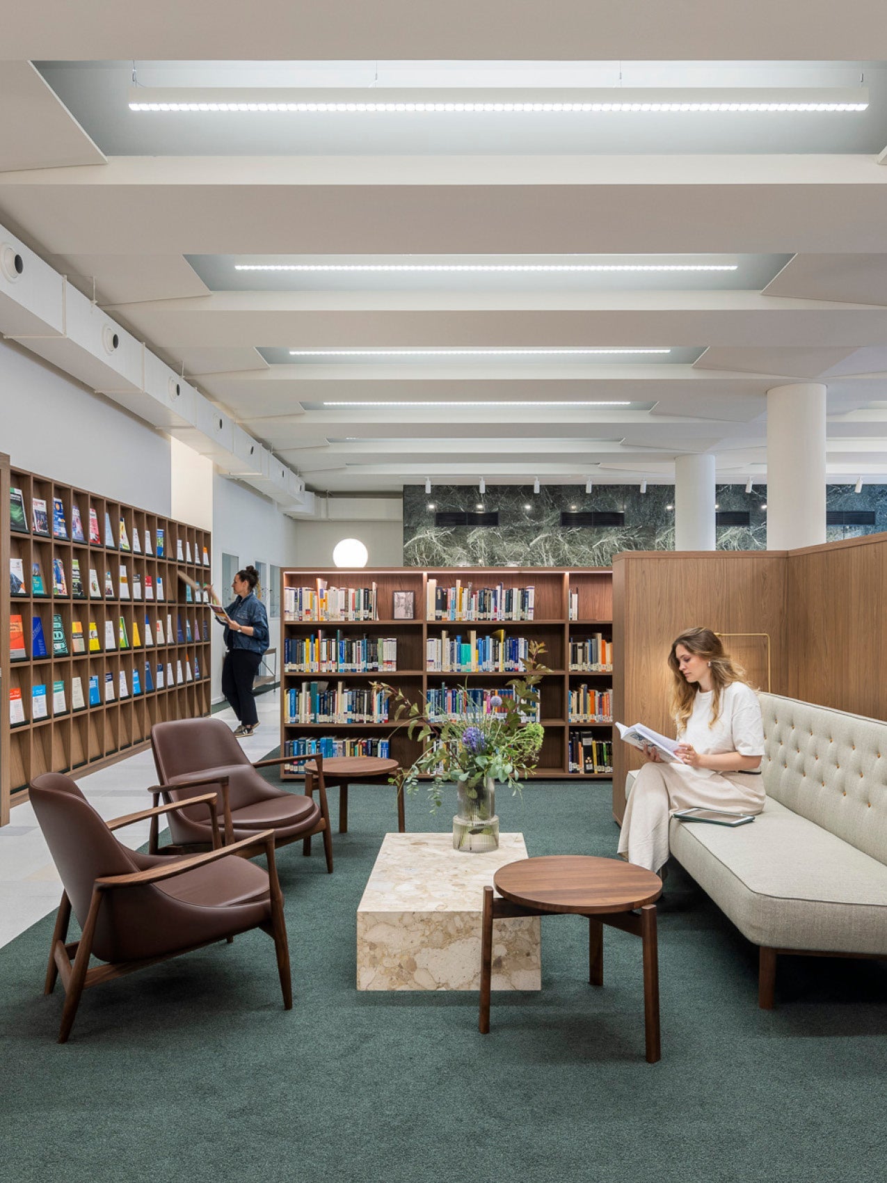 Library lounge with green carpet, walnut bookshelves, marble coffee table, brown leather chairs and a woman reading on a