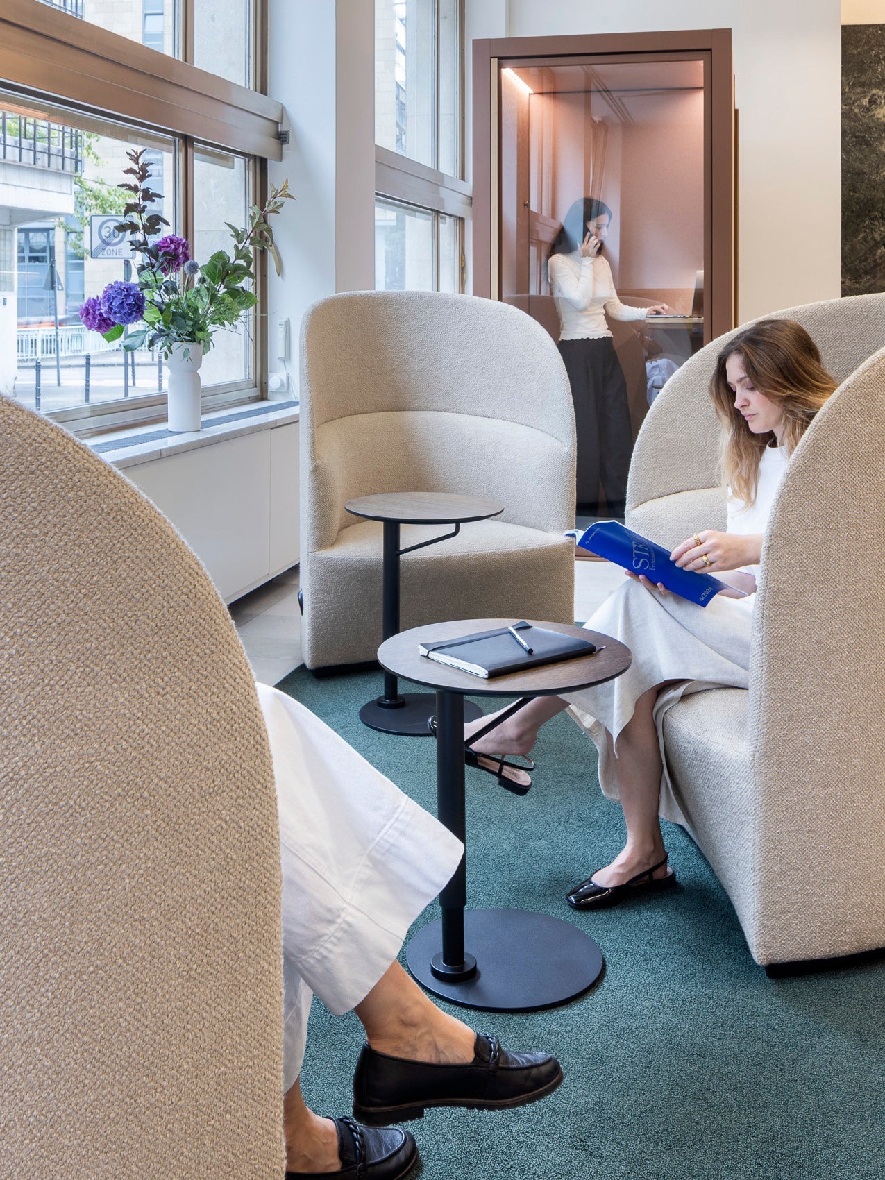 Modern beige boucle high-back lounge chairs with round black side tables in a carpeted lounge; person reading and phone