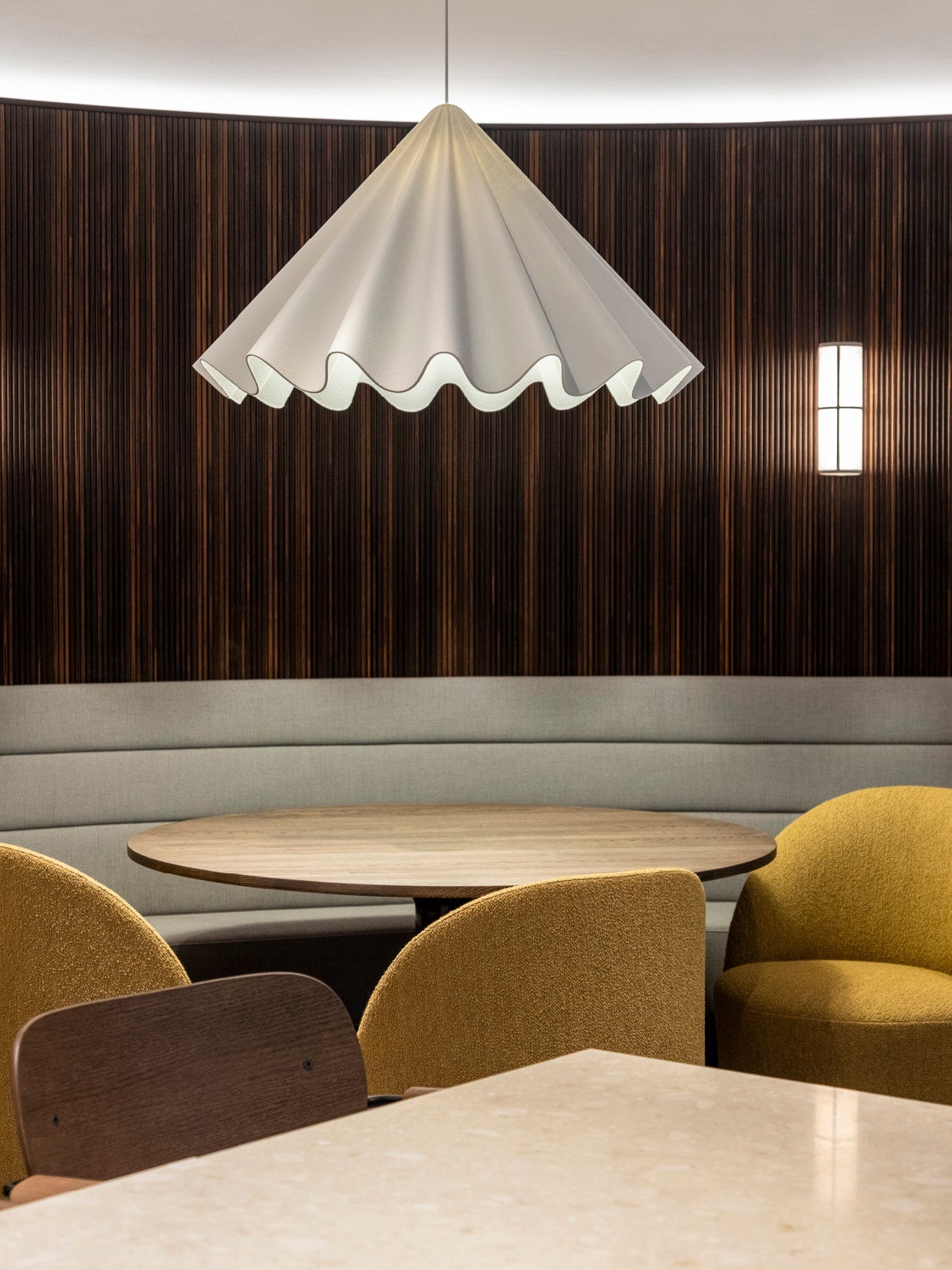White scalloped fabric pendant lamp above round wood table, grey banquette and mustard upholstered chairs against dark wood