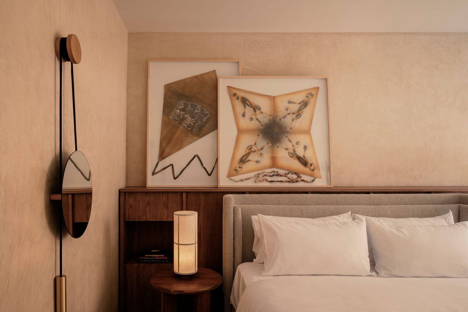 Minimal bedroom corner with beige textured walls, framed textile art, wooden headboard, round mirror and soft linen pillows