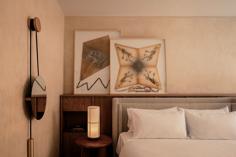 Minimal bedroom corner with beige textured walls, framed textile art, wooden headboard, round mirror and soft linen pillows