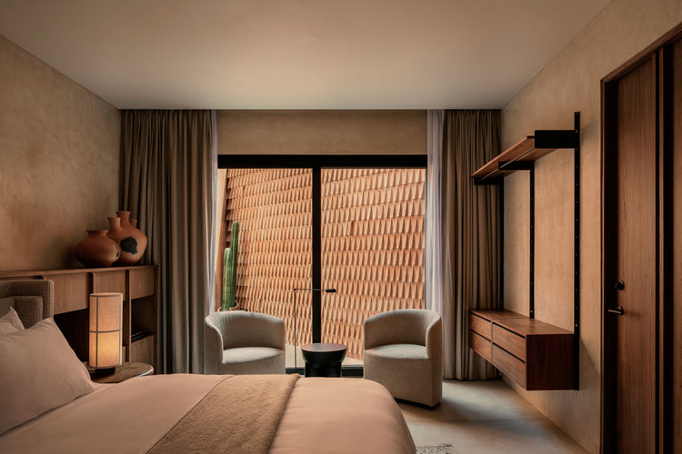 Minimal hotel bedroom with beige plaster walls, wooden shelving, two rounded beige armchairs and textured terracotta screen