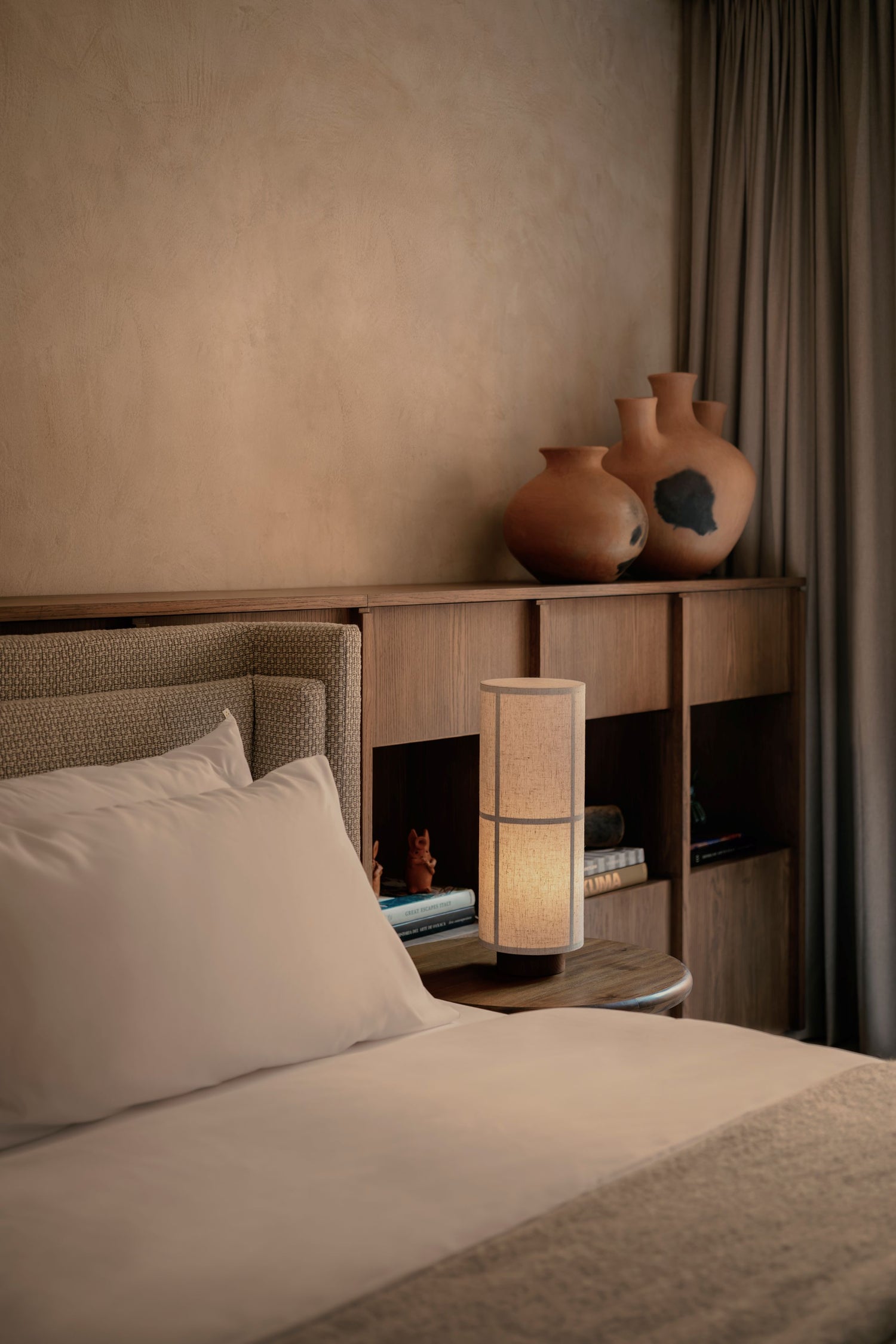 Minimal bedroom corner with neutral linen bed, wooden bedside shelf, tall fabric table lamp and two terracotta vases.
