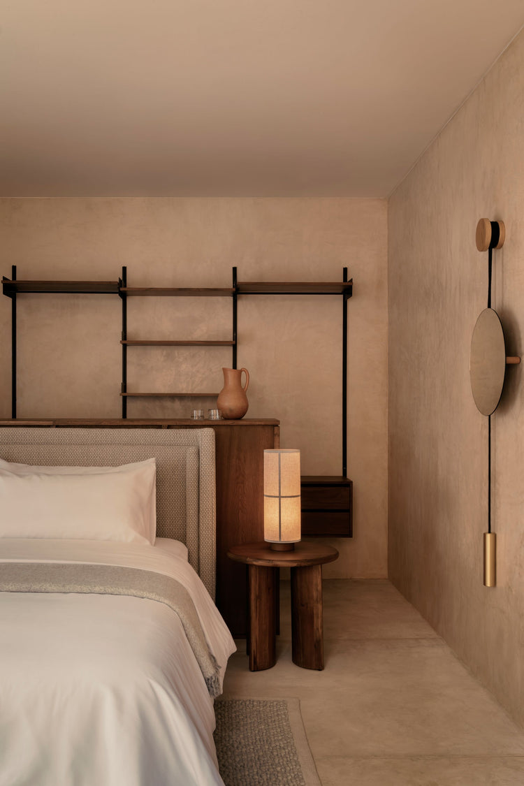 Minimal bedroom corner with beige upholstered bed, wooden bedside table and glowing fabric table lamp, wall mirror and