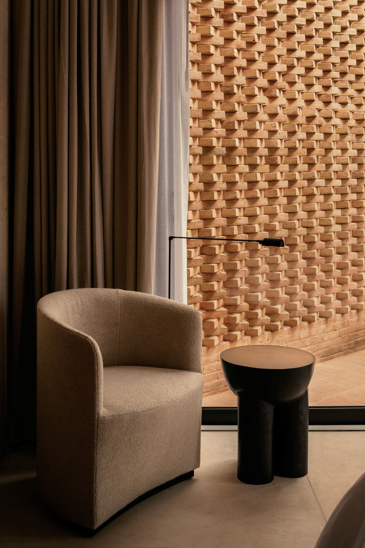 Beige upholstered curved armchair beside sheer and dark brown curtains, black sculptural side table and textured terracotta