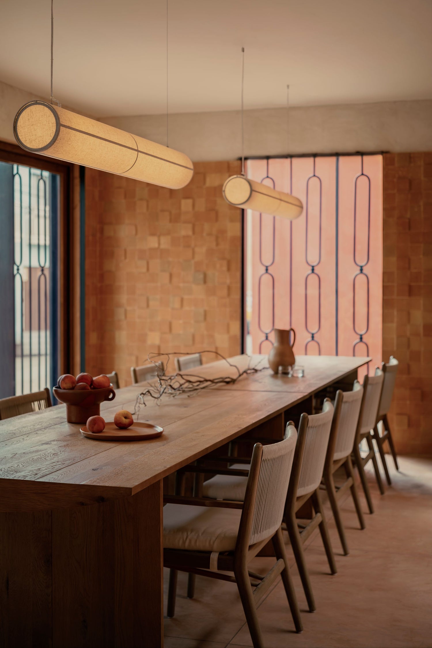 Long cylindrical beige fabric pendant lights with dark trim suspended above a wooden dining table and chairs.