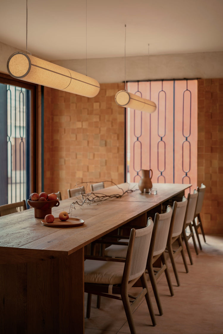 Long cylindrical beige fabric pendant lights with dark trim suspended above a wooden dining table and chairs.