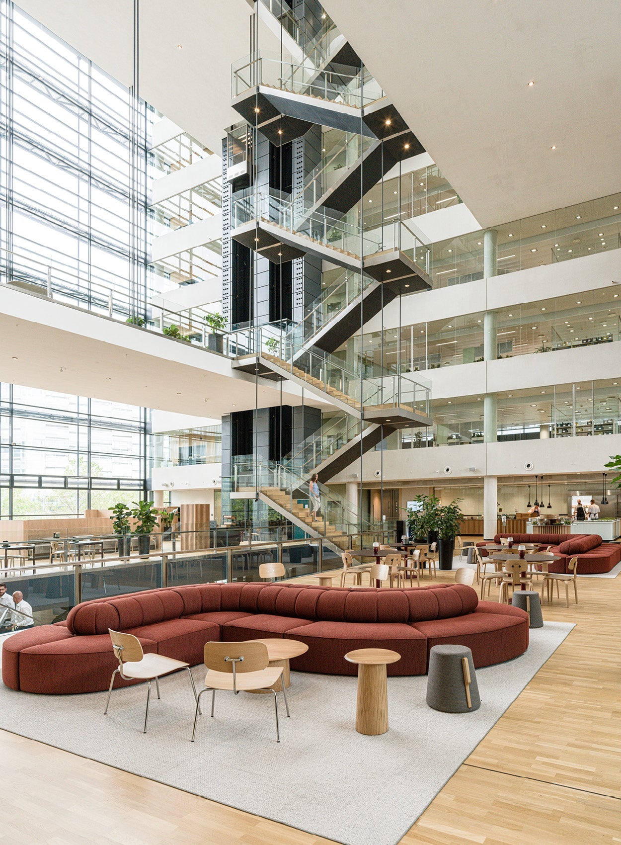 Open, multi-story atrium with glass stair tower and elevators, warm wood floors, curved rust-red modular sofa and light wood