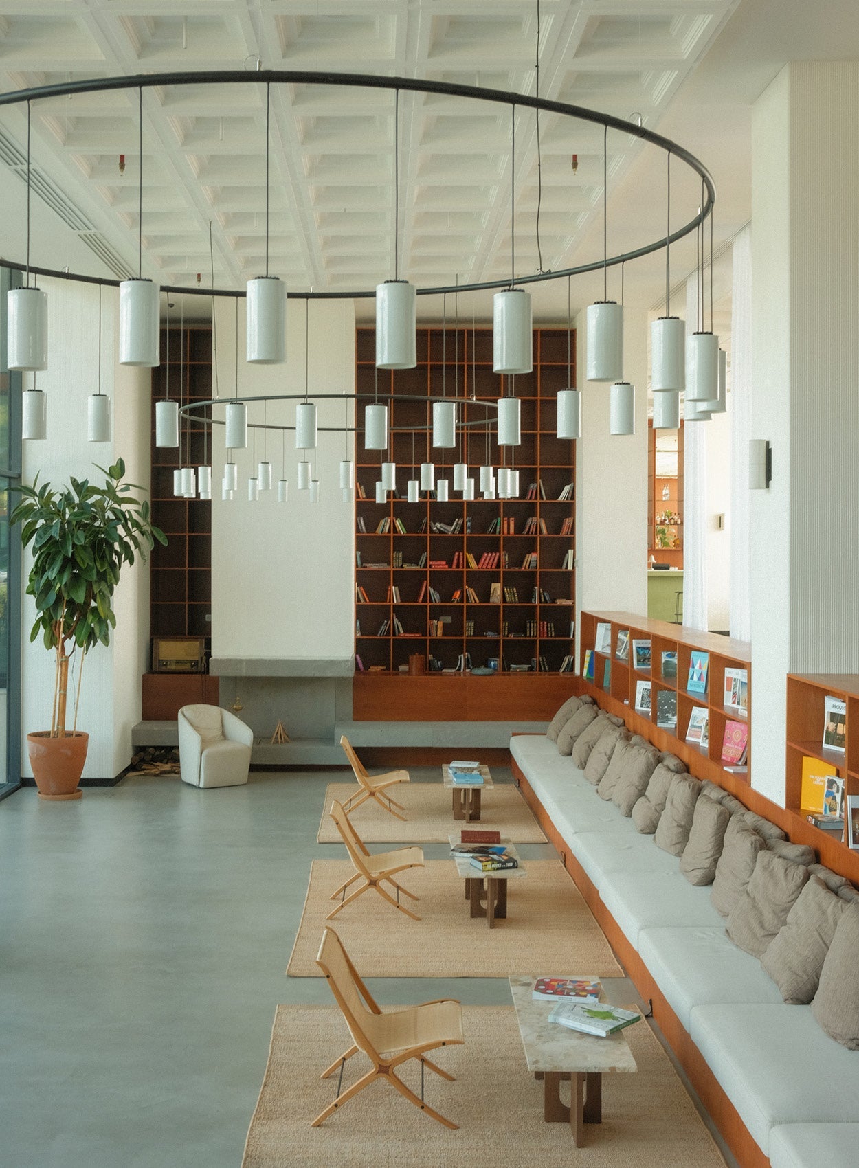Spacious lounge with long light-gray sofa, brown cushions, wooden shelving, pendant cylinder lights on black circular frames.
