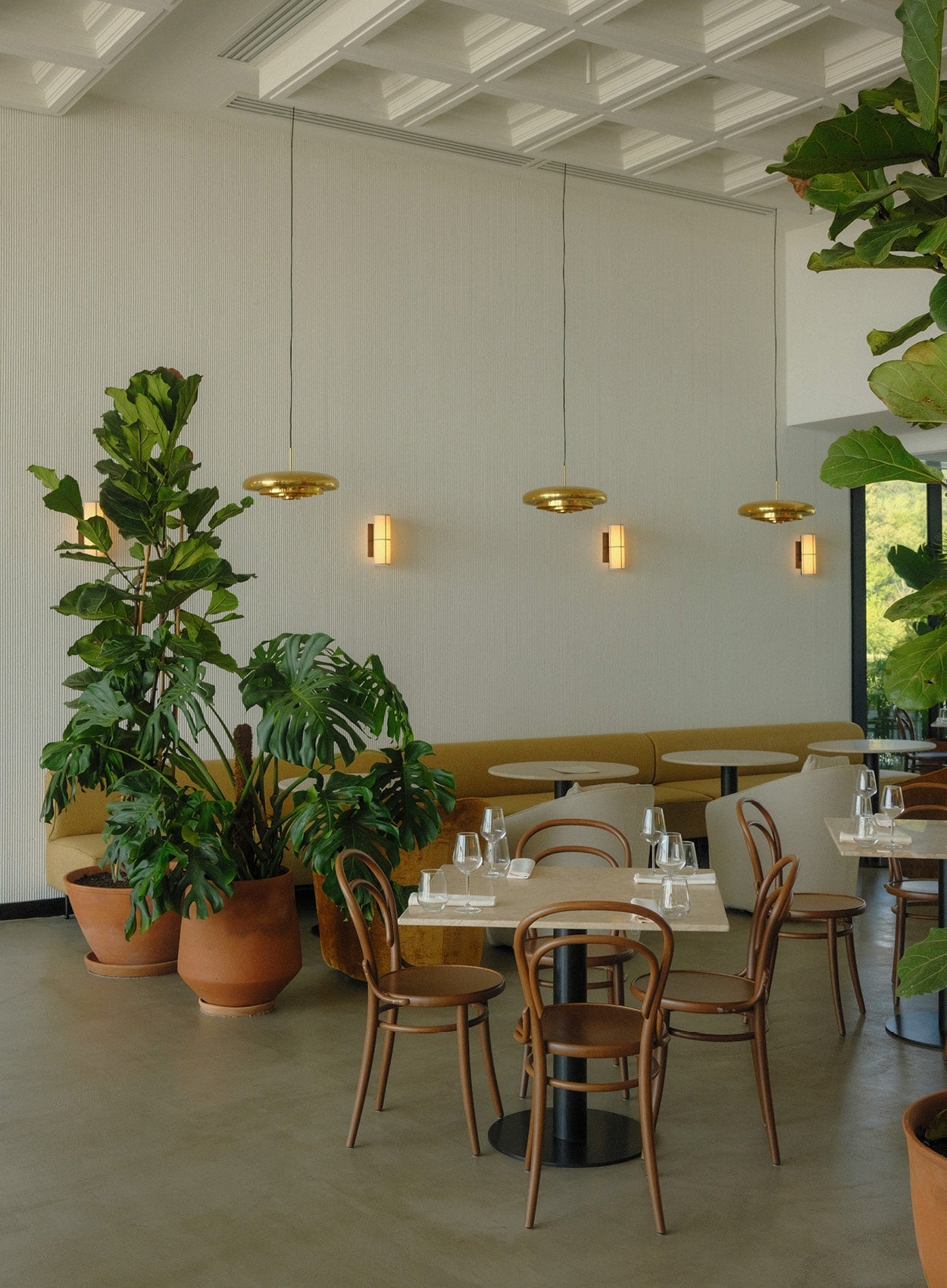 Minimal dining area with round wooden chairs, beige banquette seating, terracotta potted green plants and three brass