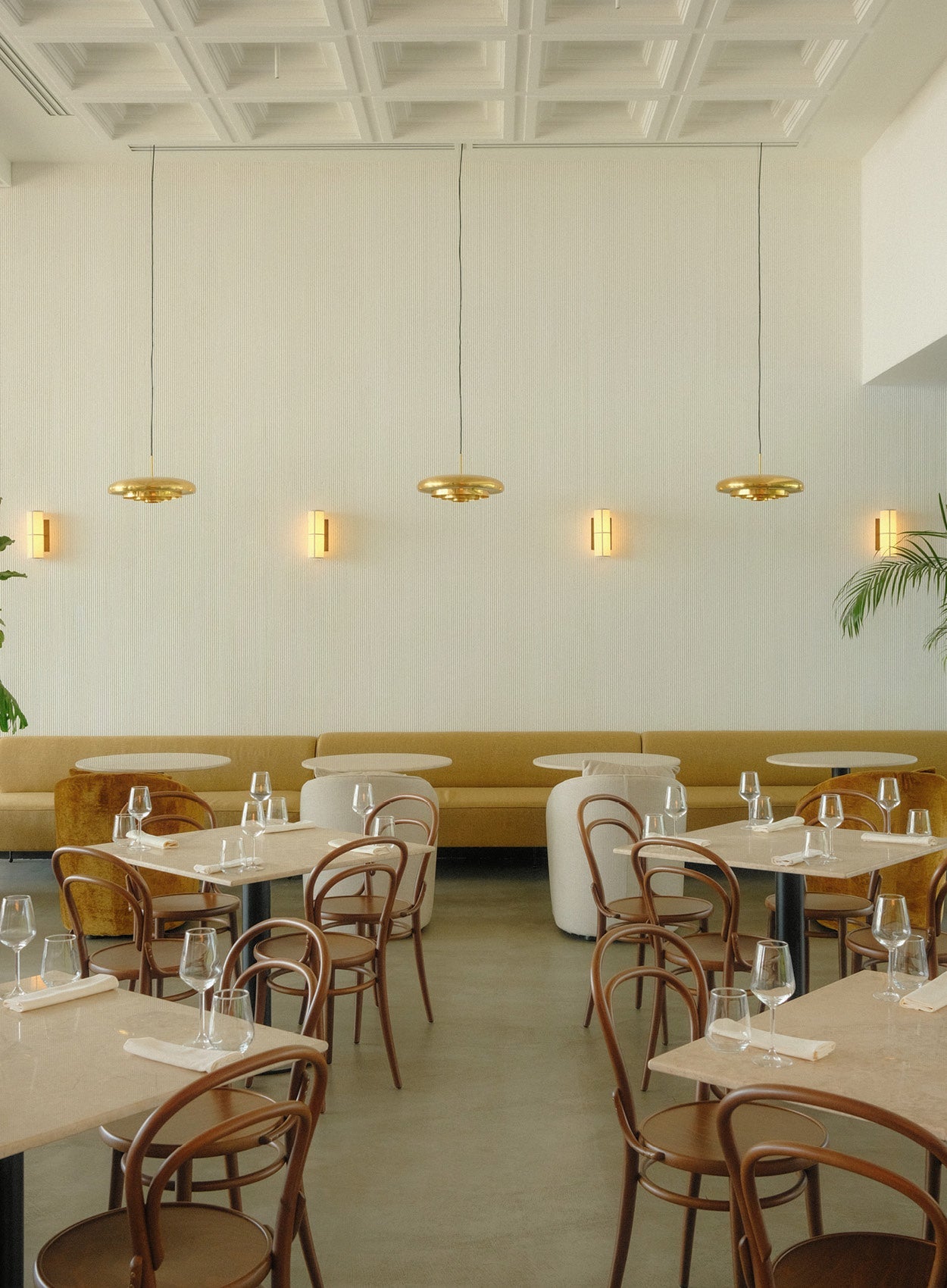 Minimal dining room with tan banquette, round marble tables, bentwood chairs and three brass disc pendant lights over seating