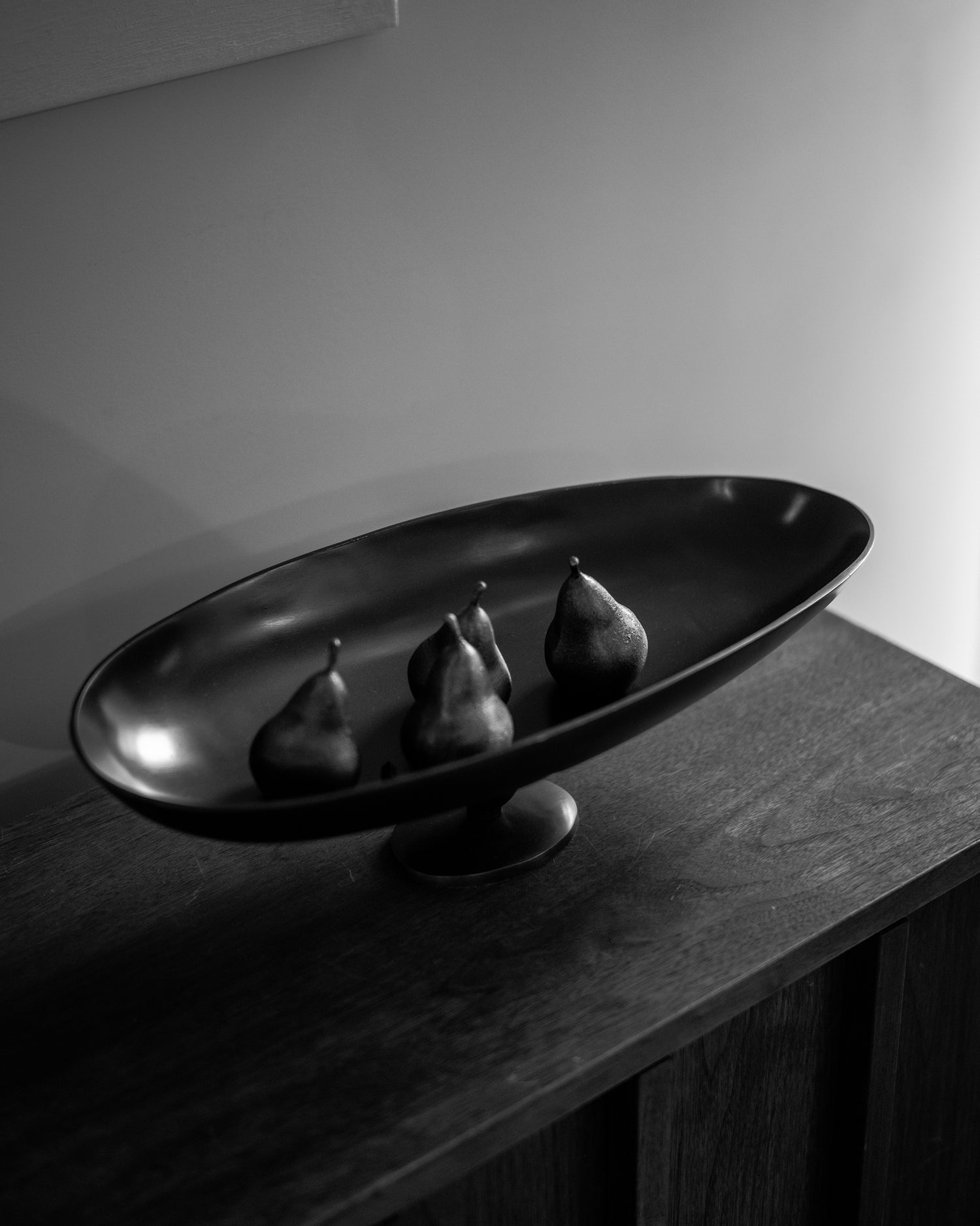 Black matte oval pedestal bowl with five dark pears arranged on a wooden sideboard, soft side lighting.