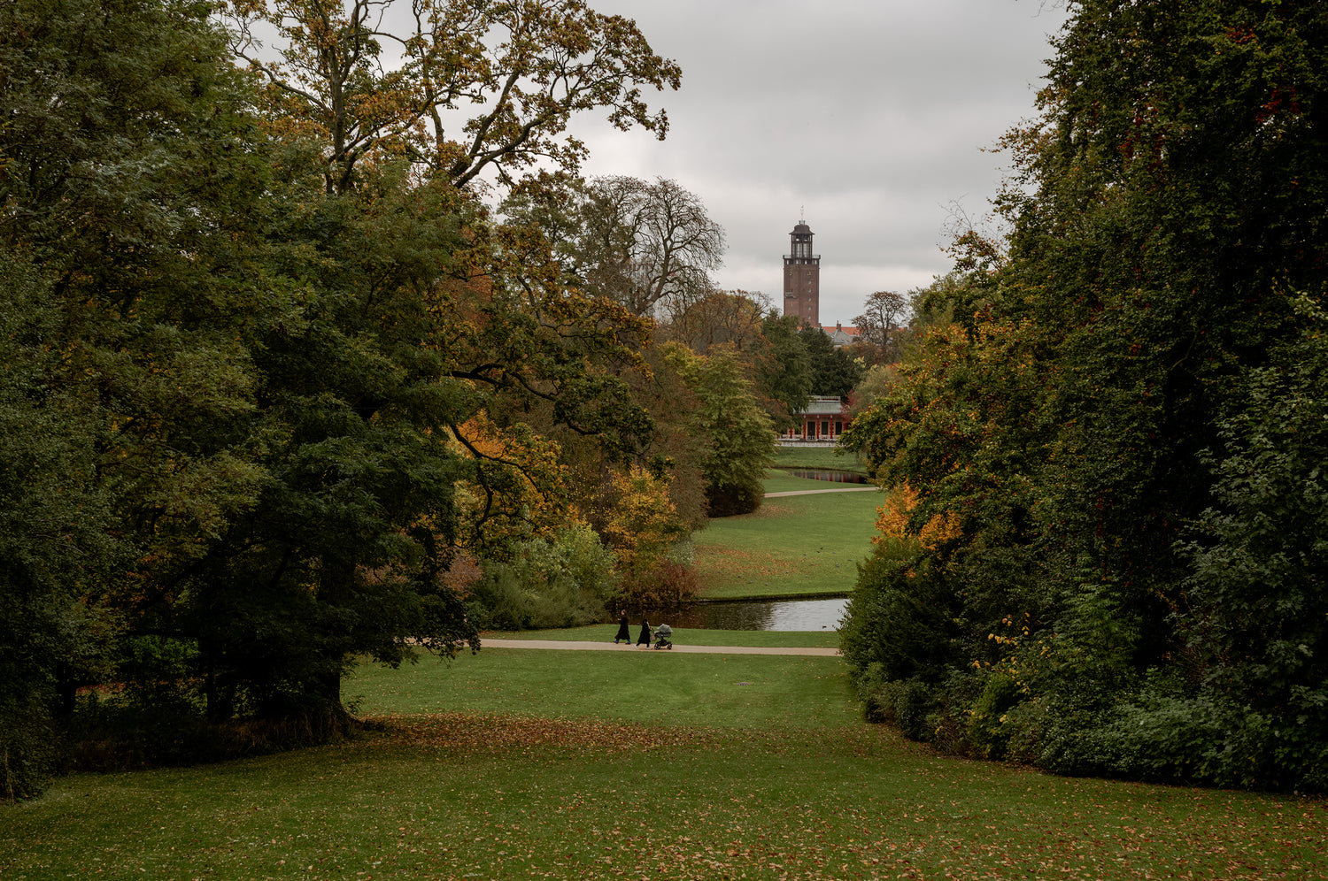 Autumn park with green lawn, large leafy trees framing a distant brick tower; two people push a stroller on path