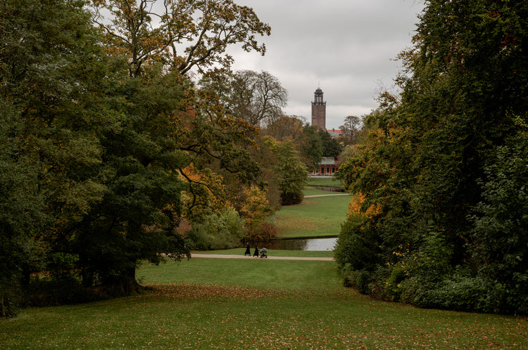 Autumn park with green lawn, large leafy trees framing a distant brick tower; two people push a stroller on path