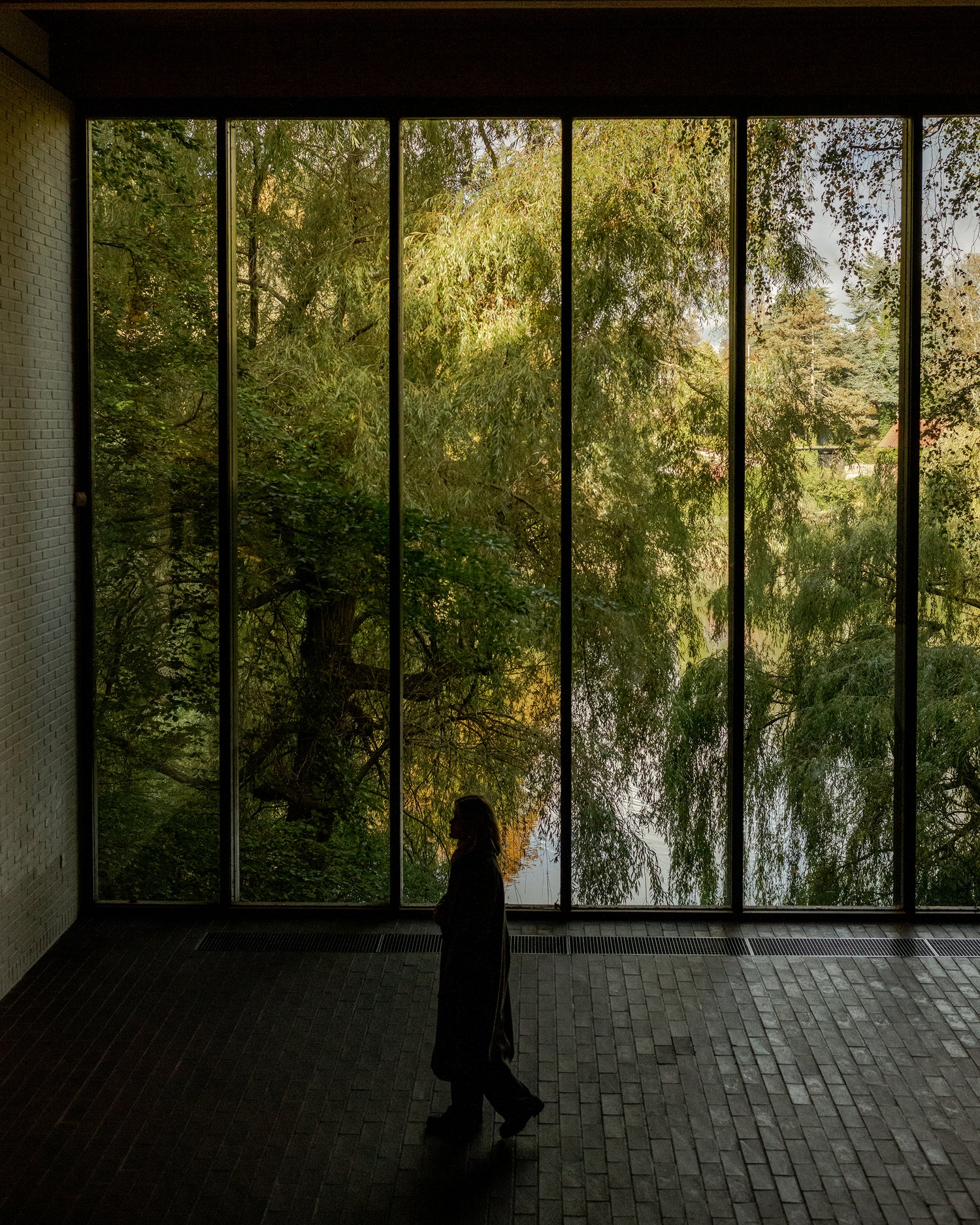 Tall floor-to-ceiling black metal-framed windows showing dense green willow trees and a silhouetted person walking inside