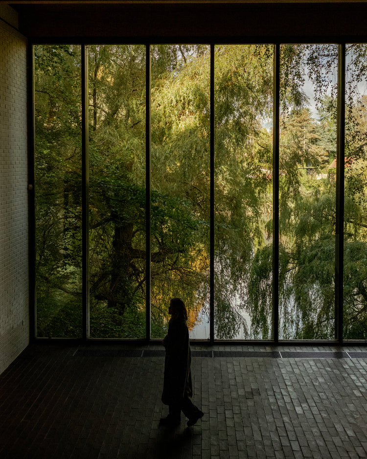 Tall floor-to-ceiling black metal-framed windows showing dense green willow trees and a silhouetted person walking inside