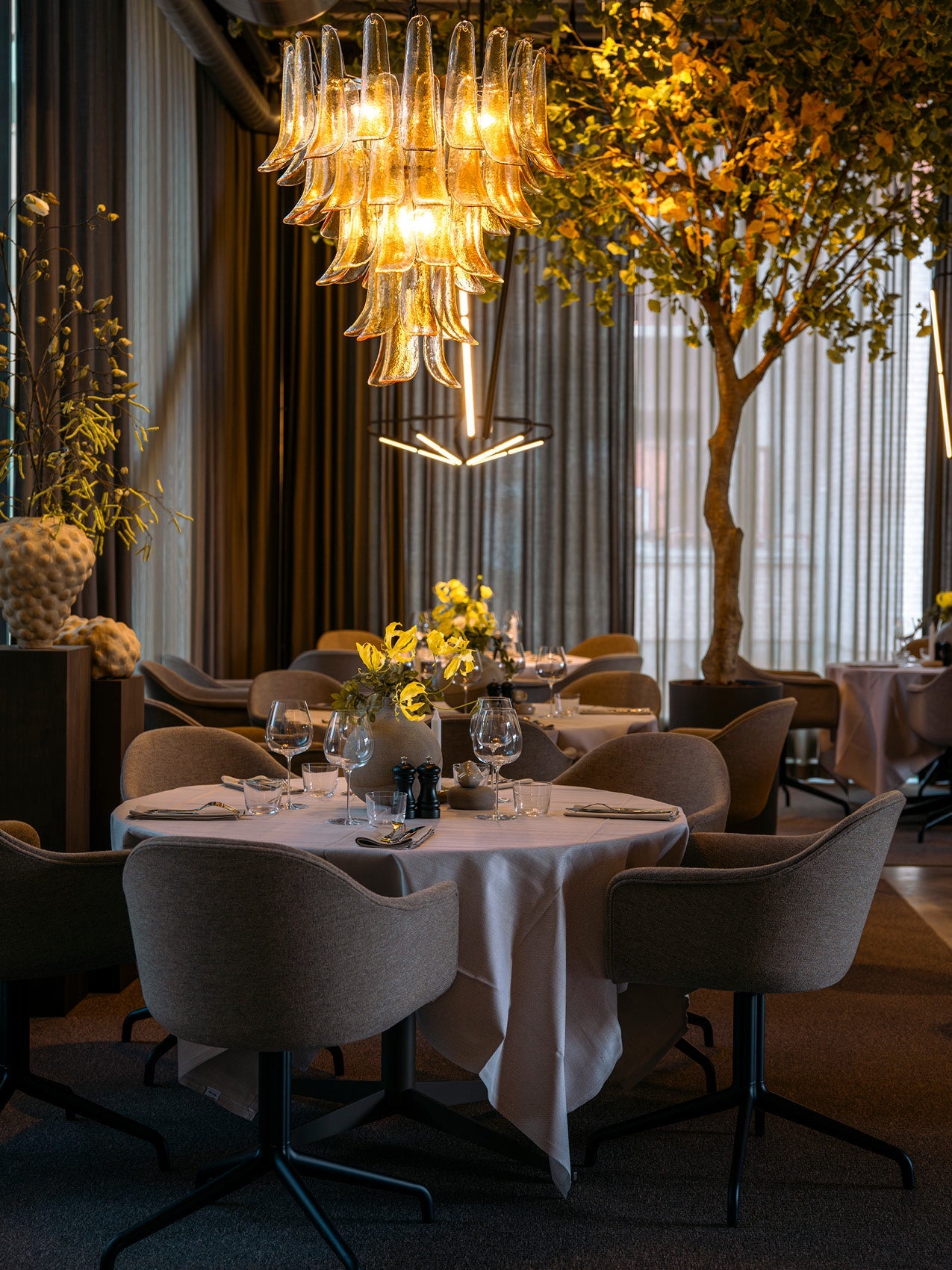 Dining table set with grey upholstered chairs, white tablecloth and glassware beneath a layered amber glass chandelier.