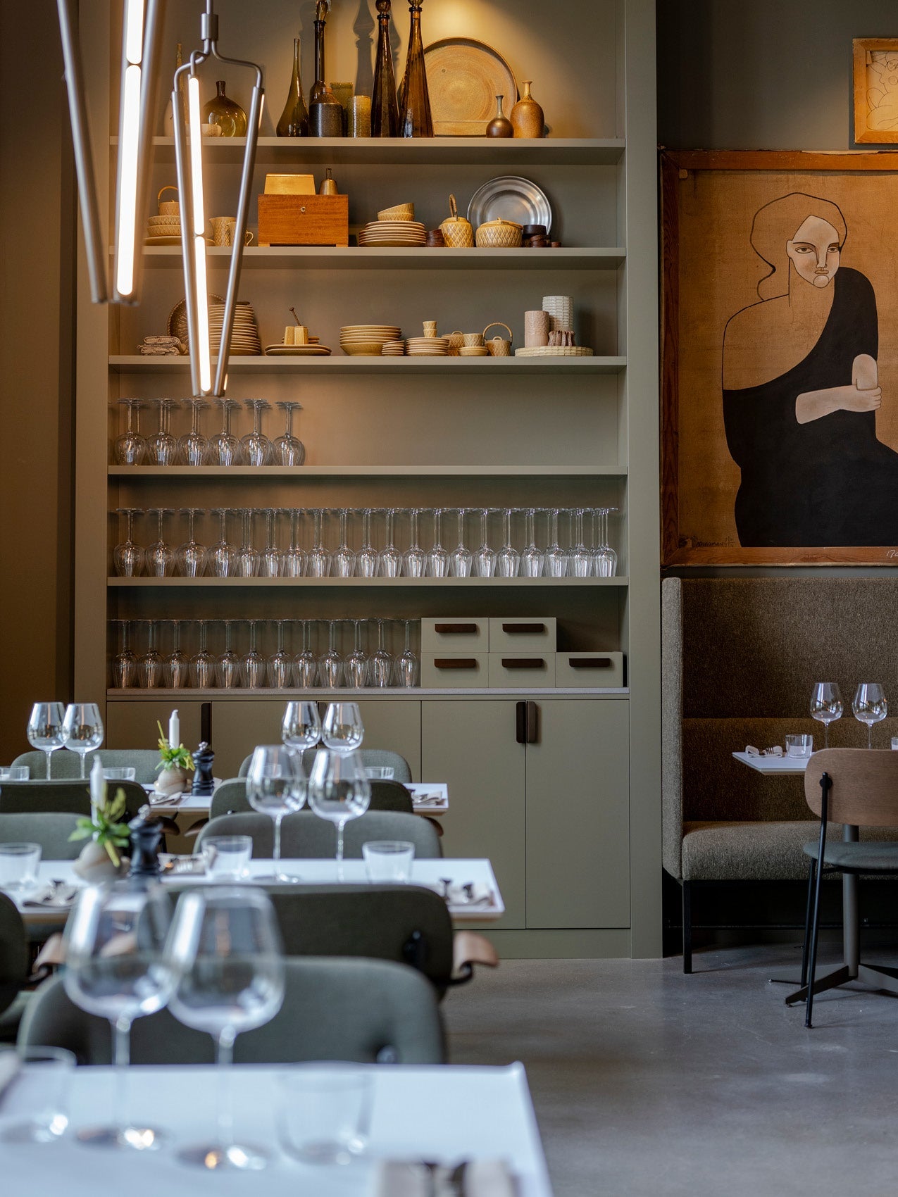 Dining room scene with olive-green shelving holding stacked plates, glasses and wooden ceramics, pendant lights and set