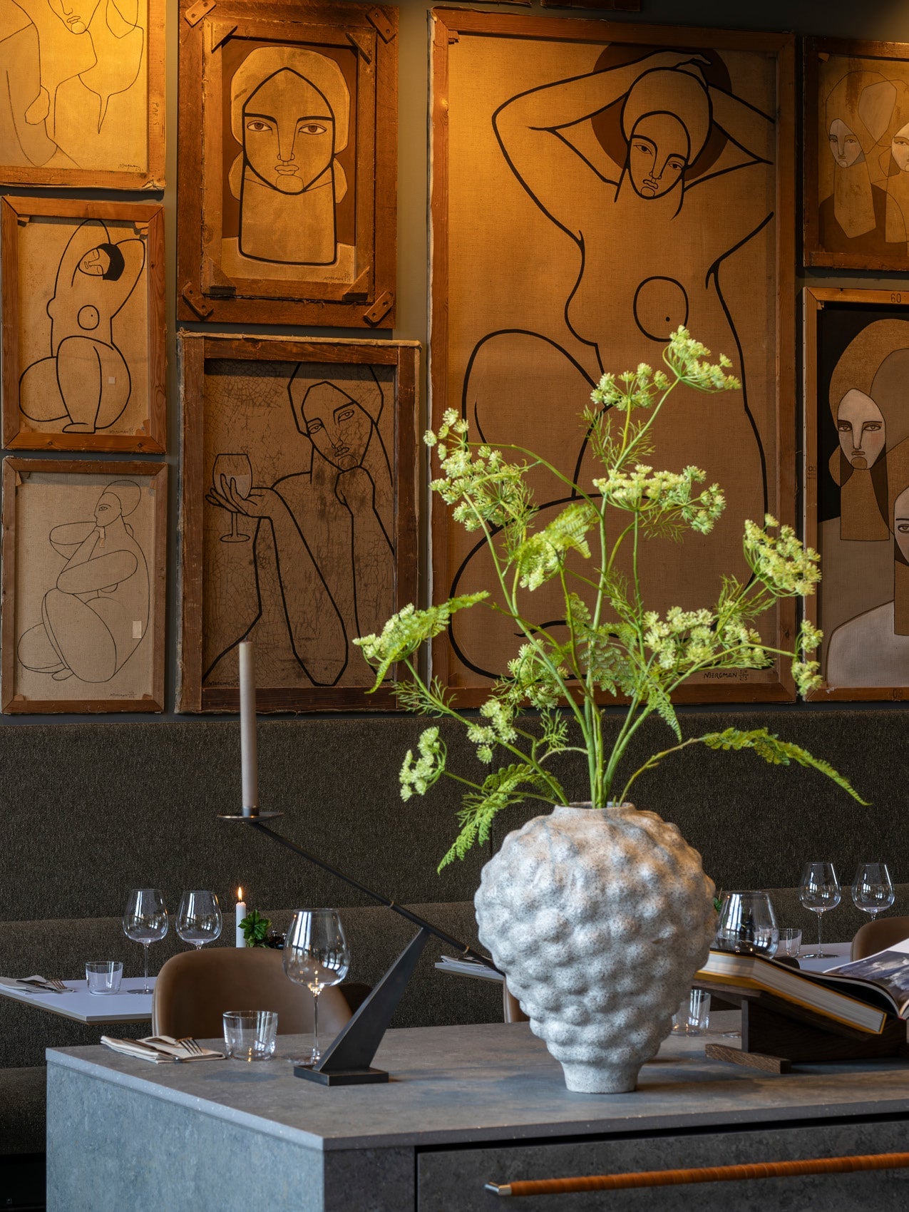 Textured white ceramic vase with green wildflowers on a dining table, backdrop of framed minimalist line-drawings in warm