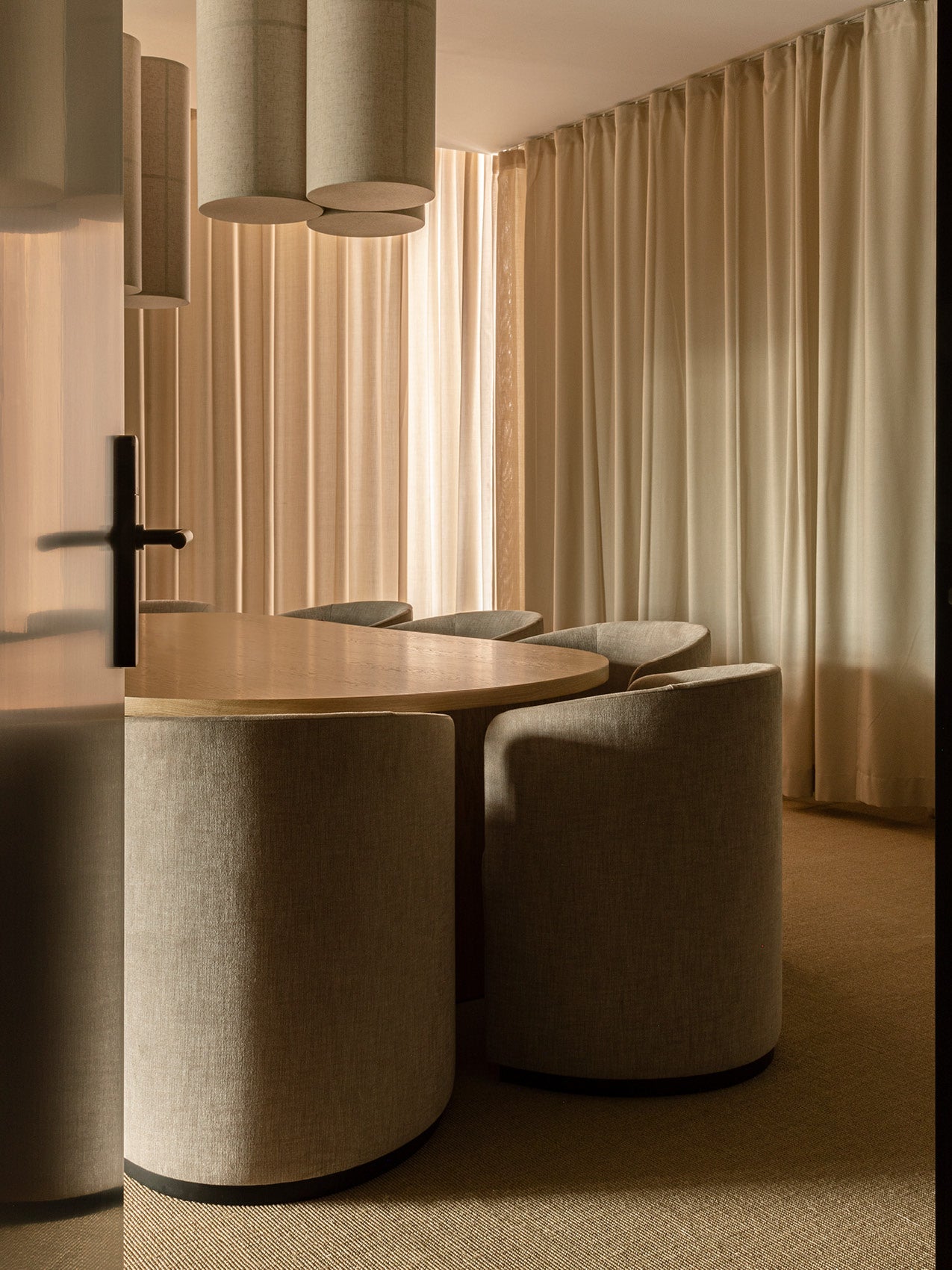 Rounded beige upholstered dining chairs around an oak oval table with beige curtains and cylindrical fabric pendant lights