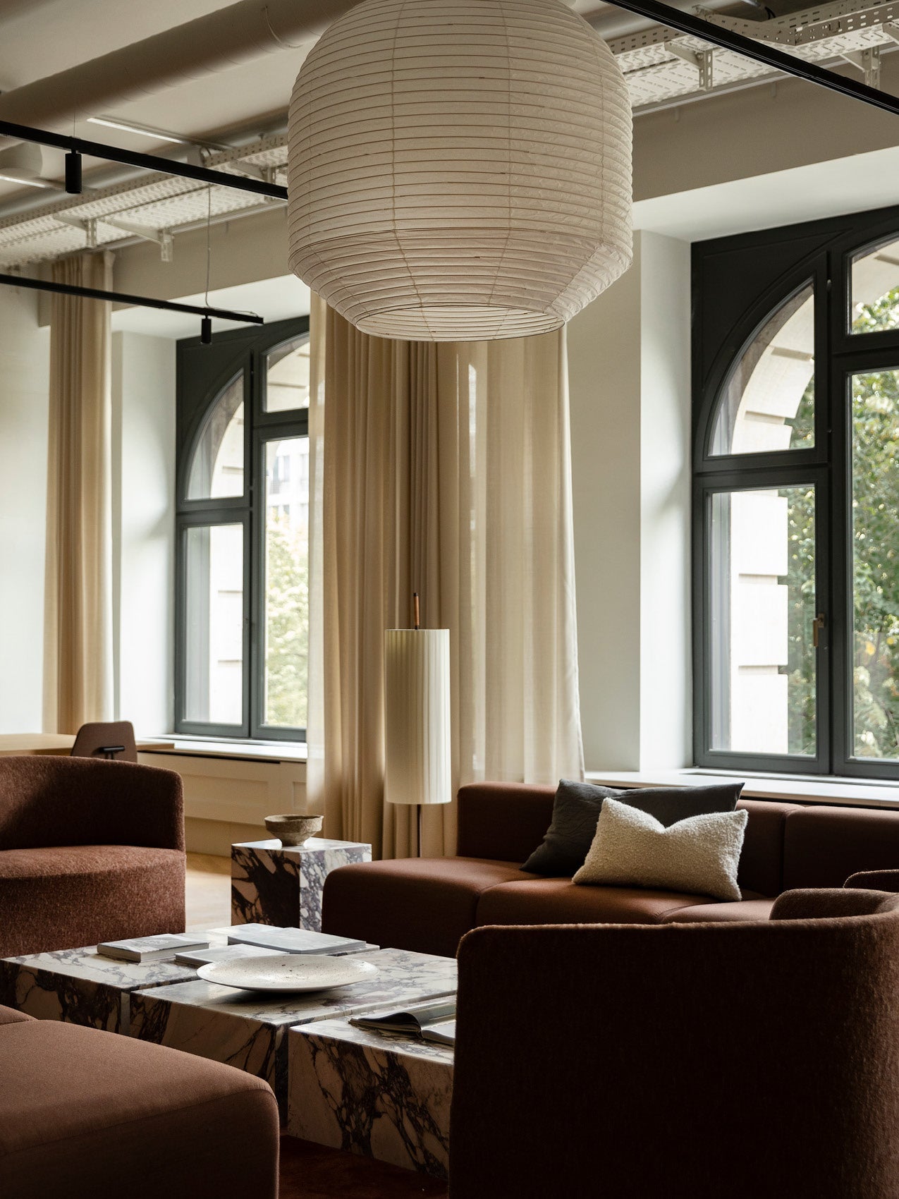 Warm living room with large white paper pendant lamp, brown upholstered sofas, marble coffee tables and tall cream curtains.