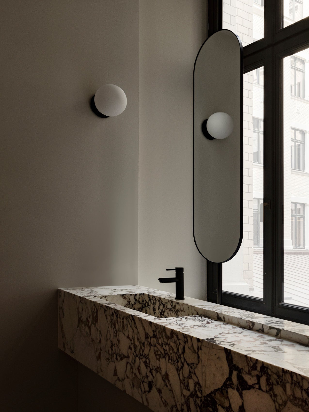 Marble sink with dark veining, matte black faucet, oval frameless mirror and round frosted wall sconce by window