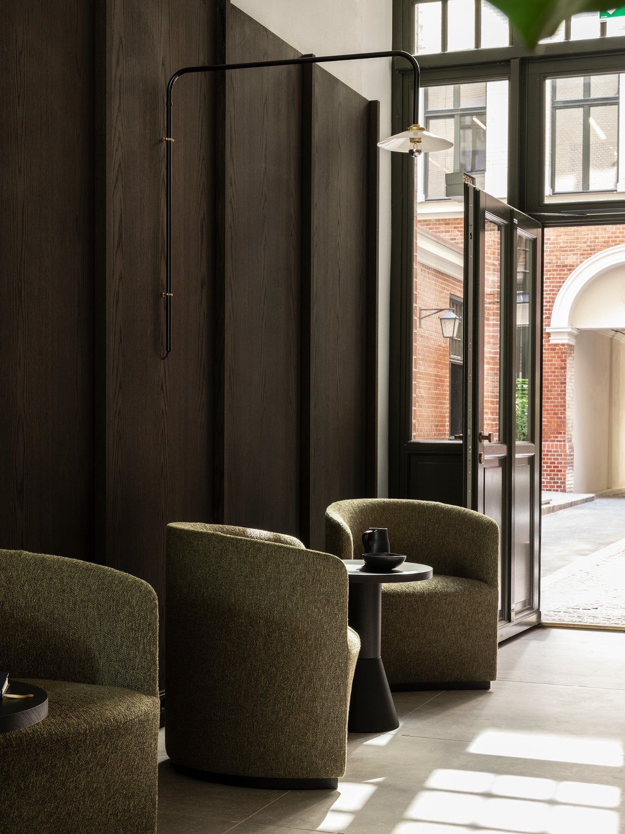 Pair of olive-green upholstered barrel chairs with a small black side table and wall-mounted black metal swing lamp near