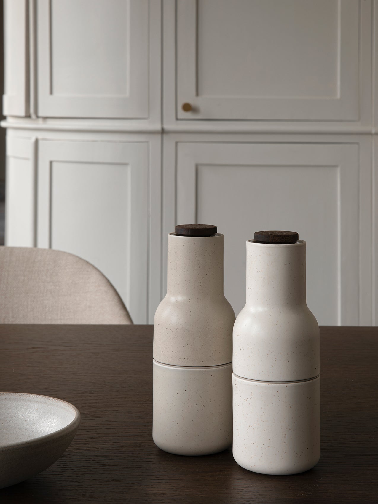 Two speckled sand ceramic Bottle Grinders with walnut lids sitting on a dark wood Adrogyne Dining Table.