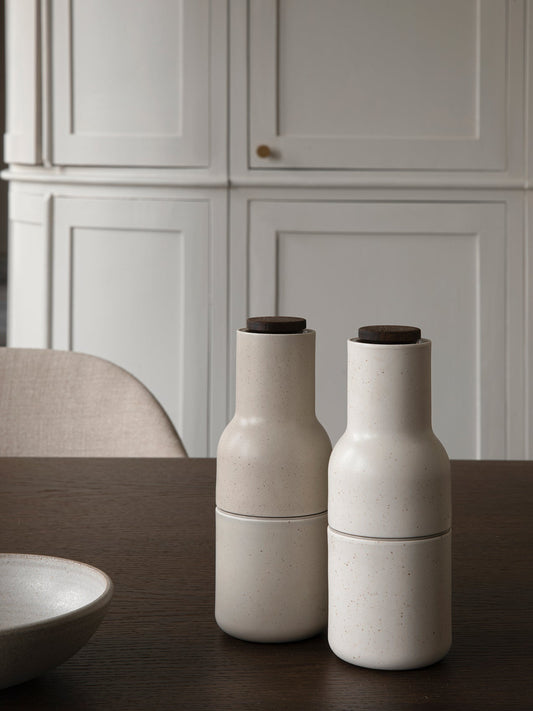 Two speckled sand ceramic Bottle Grinders with walnut lids sitting on a dark wood Adrogyne Dining Table.