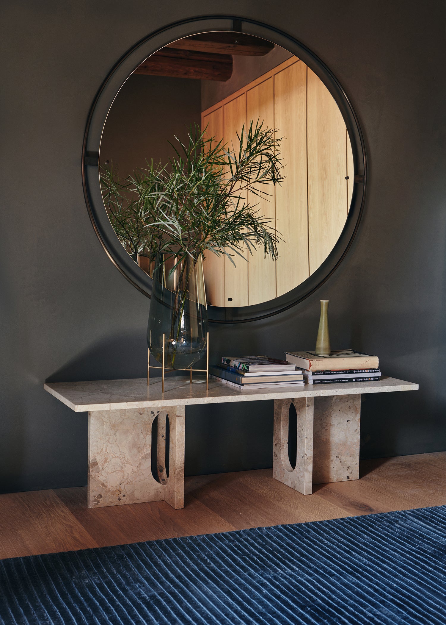 Round black-framed wall mirror reflecting wooden panels, marble console table with glass vase of green foliage and stacked