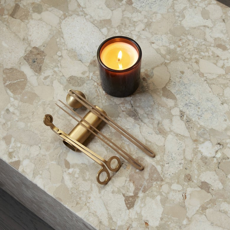 Brass candle tools (snuffer, wick trimmer, tongs) arranged beside a lit amber glass candle on beige terrazzo surface