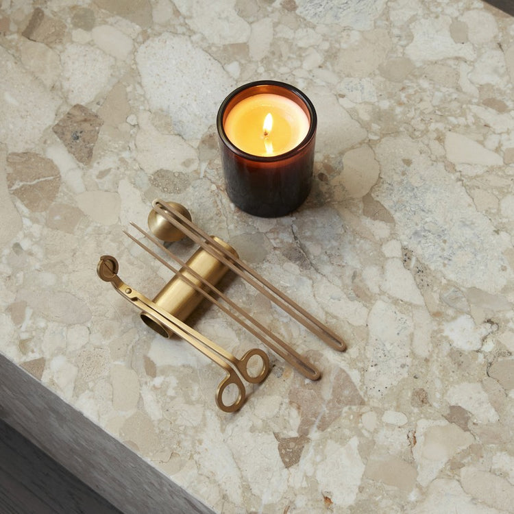 Brass candle tools (snuffer, wick trimmer, tongs) arranged beside a lit amber glass candle on beige terrazzo surface