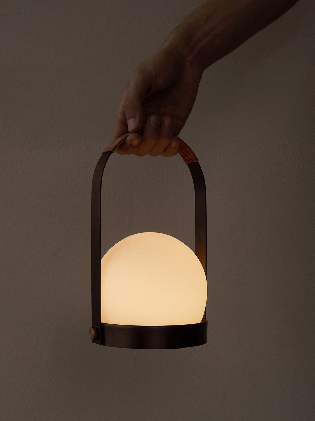 Carrie Portable Lamp, Leather Handle held by a hand, featuring a glowing opal glass shade and a dark metal frame.