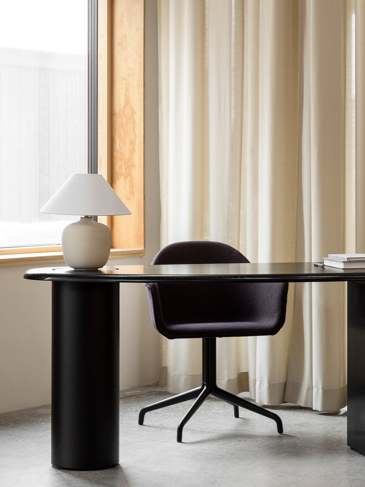 Black oval desk with cylindrical legs, dark upholstered swivel chair and beige ceramic table lamp by a wood-framed window.