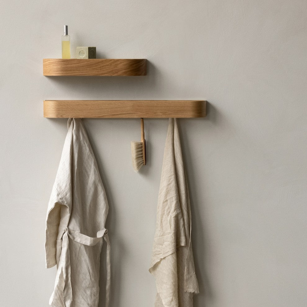Oak wall shelf and slim oak peg rail with hanging beige linen robe, towel and wooden brush on pale plaster wall.