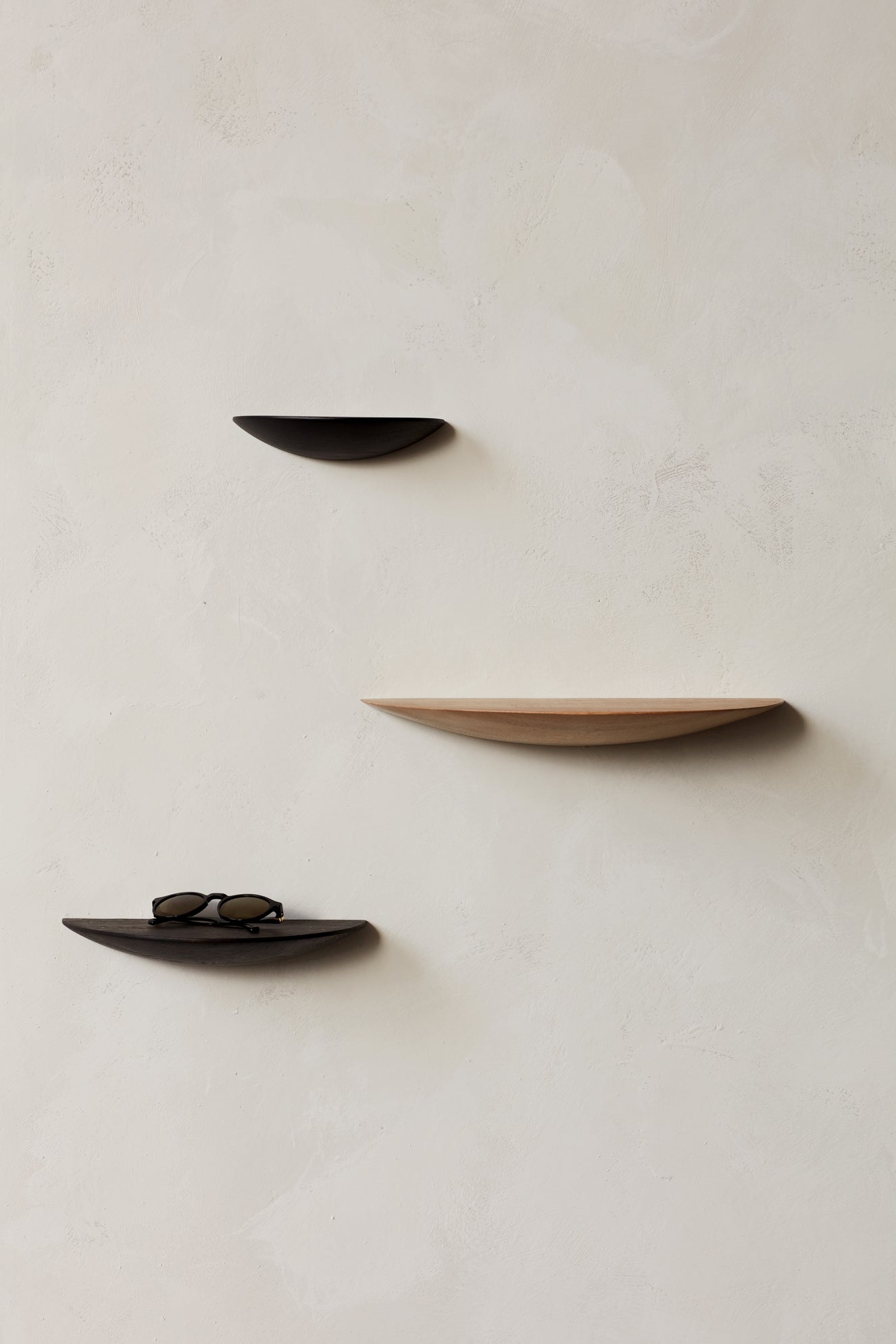 Three semicircular wall shelves in dark and natural wood mounted on a pale plaster wall; sunglasses rest on the lower shelf.